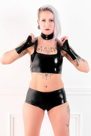A woman wearing a black latex choker, fingerless latex wrist gloves, a latex camisole top and latex boxer shorts.