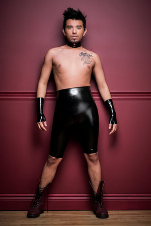 A man wearing black high waisted latex bermuda shorts, fingerless latex wrist gloves and a latex choker.