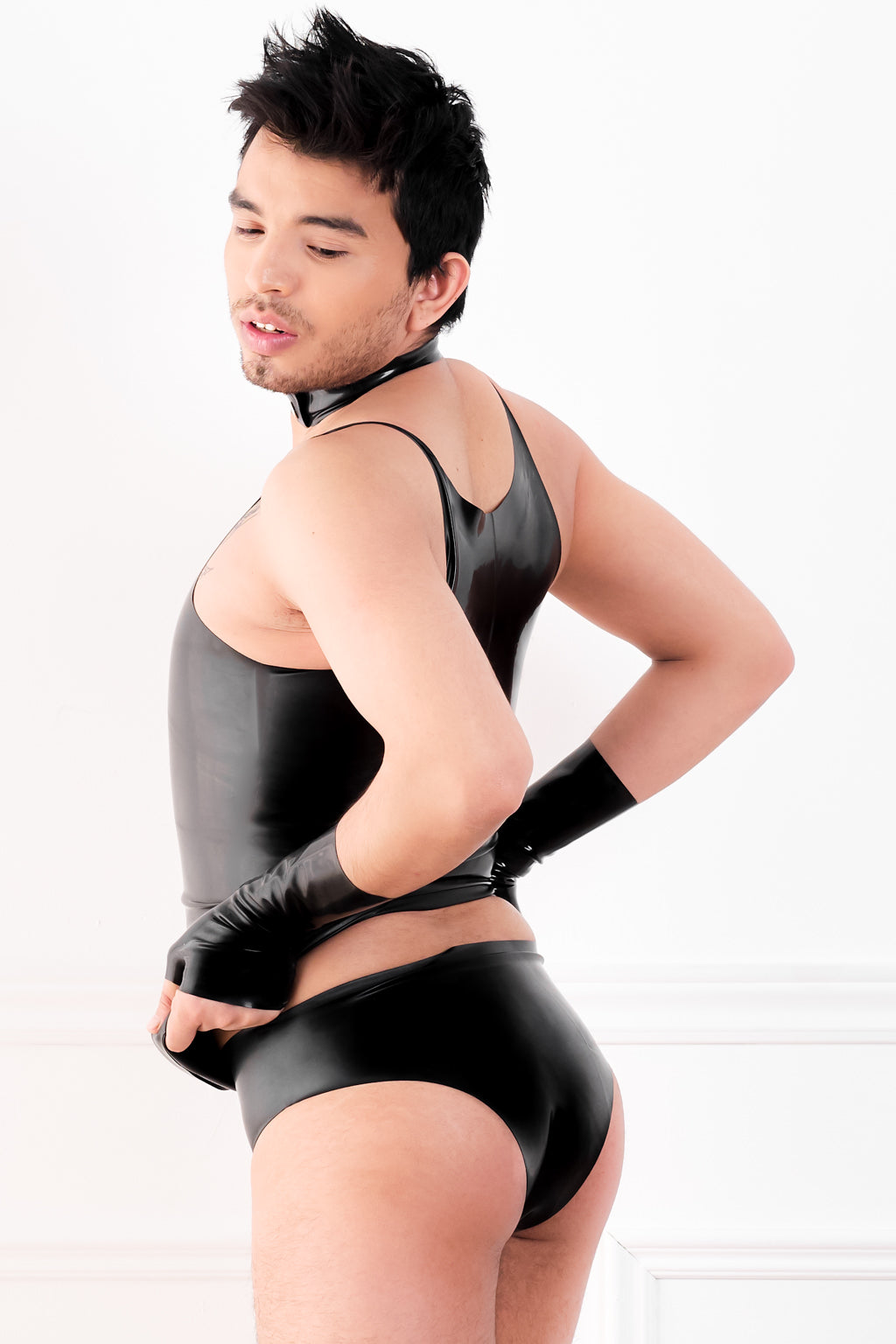 A man wearing a black latex vest, latex underwear and fingerless latex wrist gloves.